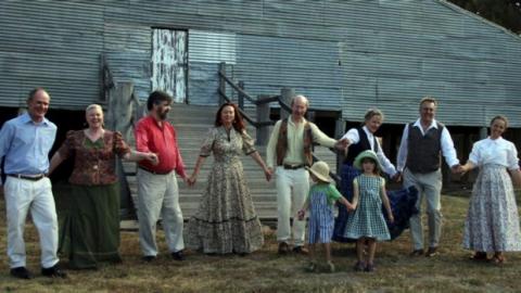 dancers in colonial dress in front of a corrugated iron woolshed